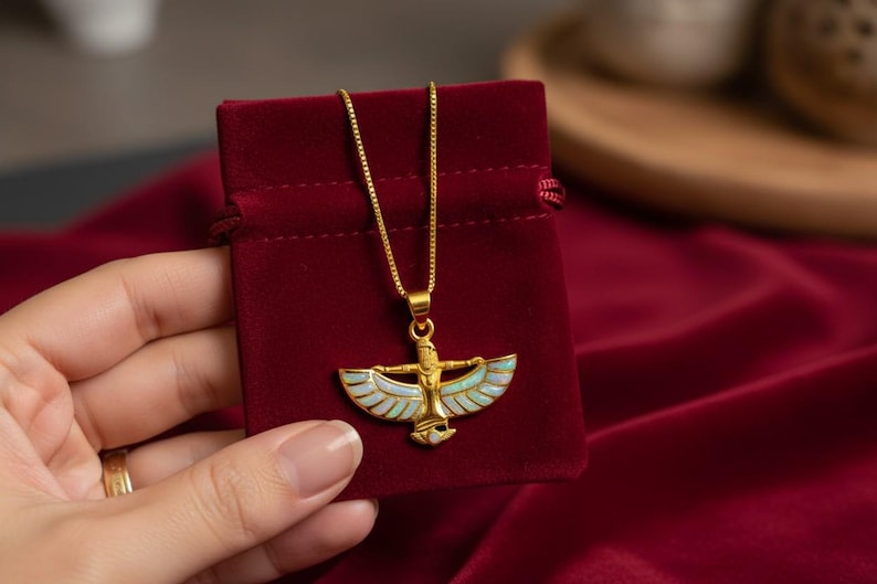 May include: A gold-colored pendant necklace featuring an Egyptian-inspired design with a figure and outstretched wings, accented with light blue and white enamel. The necklace is displayed on a red velvet pouch.