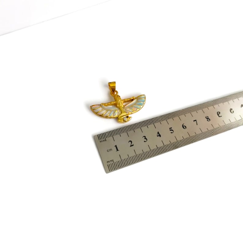 May include: Gold-colored pendant in the shape of a winged figure, with white, blue, and pink accents. The pendant is approximately 1 inch wide. The pendant is on a white surface. A ruler is next to the pendant.