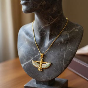 May include: A gold-colored pendant necklace with an Egyptian-inspired design, featuring a central figure with outstretched wings and colorful enamel accents. The necklace is displayed on a gray marble bust, showcasing the jewelry's intricate details.