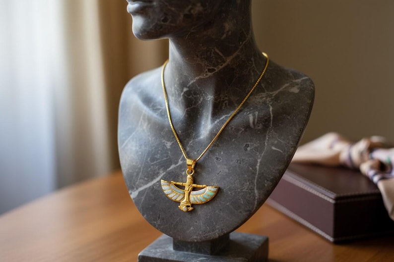 May include: A gold-colored pendant necklace with an Egyptian-inspired design, featuring a central figure with outstretched wings and colorful enamel accents. The necklace is displayed on a gray marble bust, showcasing the jewelry's intricate details.