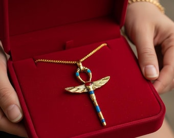 Puede incluir: Un collar con colgante Ankh dorado con detalles azules y rojos, presentado en una caja de joyería de terciopelo rojo. El Ankh, símbolo de vida, es una cruz con un lazo, a menudo vinculado al antiguo Egipto.