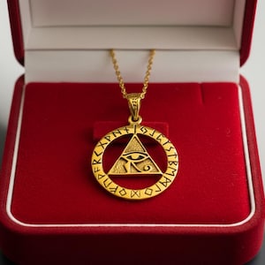May include: A gold-coloured pendant necklace with an eye within a triangle, surrounded by runes. The necklace is presented in a red velvet jewellery box. The pendant has a detailed design and is suitable for symbolic wear.