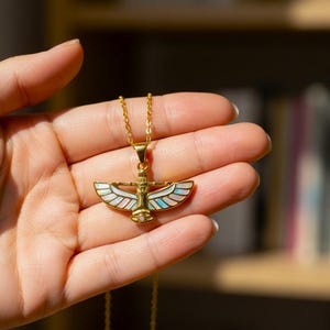 May include: A gold-colored necklace with a pendant featuring an Egyptian-inspired design. The pendant has a central figure with outstretched wings, adorned with iridescent mother-of-pearl and turquoise accents. The necklace is held in a hand.