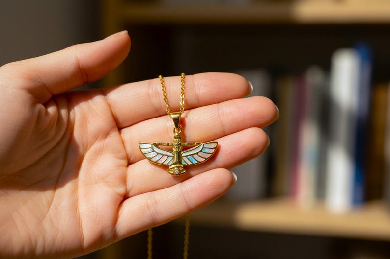 May include: A gold-colored necklace with a pendant featuring an Egyptian-inspired design. The pendant has a central figure with outstretched wings, adorned with iridescent mother-of-pearl and turquoise accents. The necklace is held in a hand.