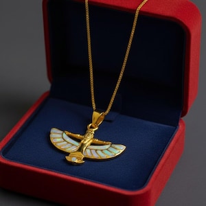 May include: Gold-toned pendant necklace featuring an Egyptian-inspired design, presented in a red velvet jewelry box with a blue interior. The pendant showcases a stylized figure with outstretched wings, accented with iridescent white and gold details.