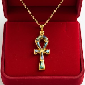 Könnte beinhalten: Eine goldfarbene Ankh-Anhänger-Halskette, präsentiert in einer roten Samt-Schmuckschatulle. Der Anhänger hat schillernde Abalone-Muschel-Inlays. Die Halskette ist ein Symbol für Leben und Ewigkeit.