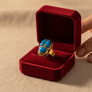 May include: A gold ring with a turquoise scarab beetle design, presented in a red velvet ring box. The ring box is open, and a hand is reaching towards it. The ring is a statement piece.