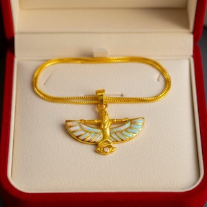 May include: A gold-colored pendant necklace featuring an Egyptian-style design with a winged figure, set in a red velvet jewelry box. The pendant has white, light blue, and gold accents. The necklace chain is also gold.