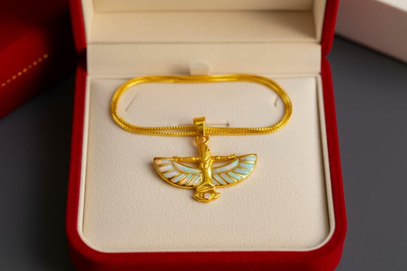 May include: A gold-colored pendant necklace featuring an Egyptian-style design with a winged figure, set in a red velvet jewelry box. The pendant has white, light blue, and gold accents. The necklace chain is also gold.