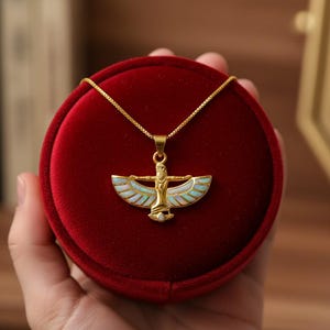 May include: Gold-toned necklace with a pendant featuring a stylized figure with outstretched wings, accented with white and light blue enamel. The pendant is displayed in a red velvet jewelry box.
