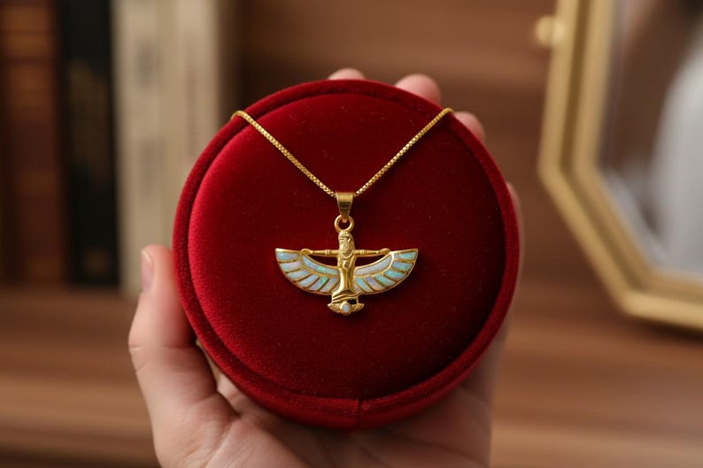 May include: Gold-toned necklace with a pendant featuring a stylized figure with outstretched wings, accented with white and light blue enamel. The pendant is displayed in a red velvet jewelry box.