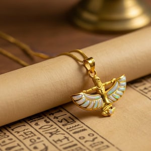 May include: Gold-toned pendant necklace featuring an Egyptian-inspired design. The pendant depicts a figure with outstretched wings, accented with white and iridescent details. The necklace rests on a scroll with hieroglyphic text.