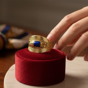 May include: A gold-colored ring with a rectangular, dark blue stone set in the center. The ring is on a red velvet display stand. The ring has an intricate, textured design.