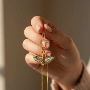 May include: A gold-colored pendant necklace featuring an Egyptian-inspired design. The pendant has a central figure with outstretched wings, accented with iridescent and white details. The necklace is held up by a delicate gold chain.