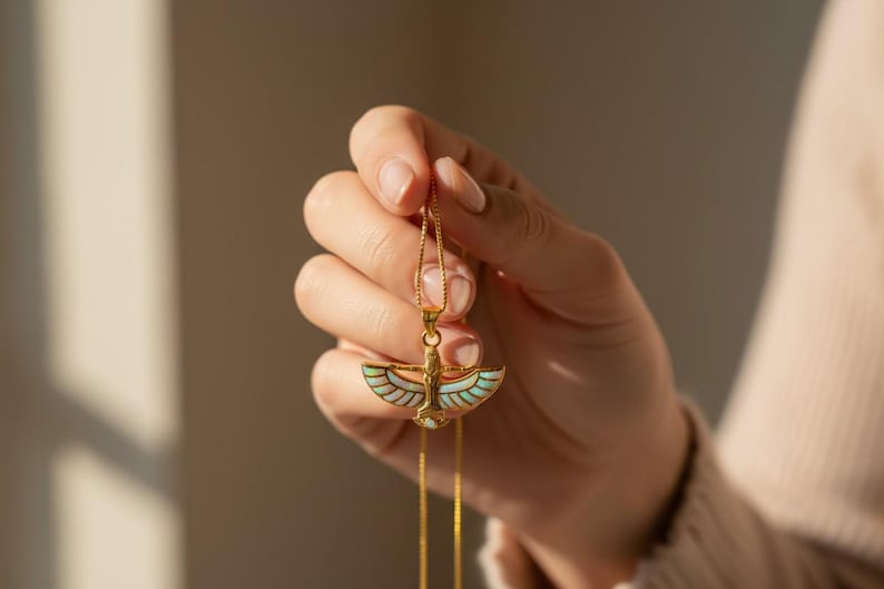 May include: A gold-colored pendant necklace featuring an Egyptian-inspired design. The pendant has a central figure with outstretched wings, accented with iridescent and white details. The necklace is held up by a delicate gold chain.