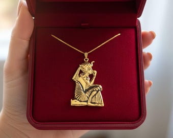 Puede incluir: Un collar con colgante dorado con un diseño de faraón egipcio, presentado en una caja de joyería de terciopelo rojo. El colgante representa a un faraón sentado con detalles intrincados. El collar es un accesorio elegante.