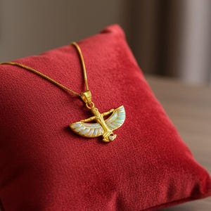 May include: A gold-colored pendant necklace featuring an Egyptian-style design with outstretched wings and a central figure. The pendant is adorned with iridescent white and light blue accents. The necklace rests on a red velvet pillow.