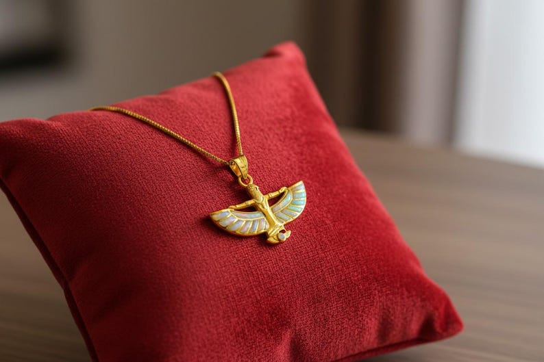 May include: A gold-colored pendant necklace featuring an Egyptian-style design with outstretched wings and a central figure. The pendant is adorned with iridescent white and light blue accents. The necklace rests on a red velvet pillow.