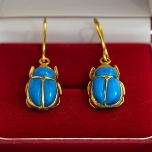 May include: A pair of gold-colored earrings shaped like scarab beetles, with bright turquoise-colored bodies. The earrings are displayed inside a red velvet jewelry box.