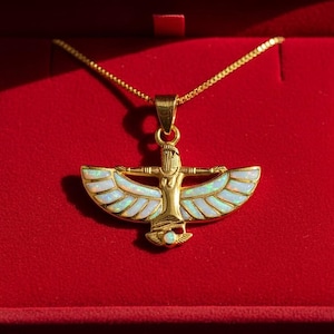 May include: Gold-toned pendant necklace featuring an Egyptian-inspired design with a central figure and outstretched wings inlaid with iridescent white stones. The necklace rests in a red velvet jewelry box.