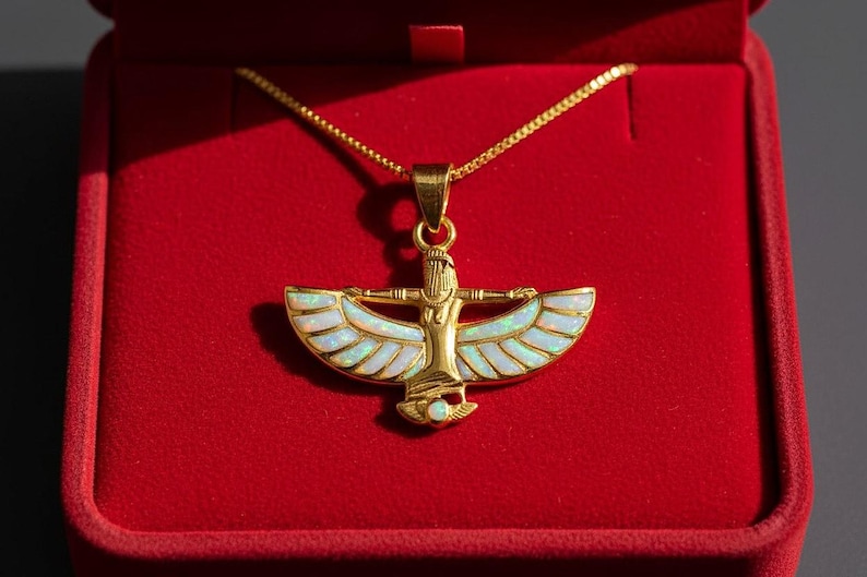 May include: Gold-toned pendant necklace featuring an Egyptian-inspired design with a central figure and outstretched wings inlaid with iridescent white stones. The necklace rests in a red velvet jewelry box.
