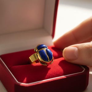 May include: A gold ring with a blue scarab beetle design, presented in a red velvet box. The ring features a detailed beetle with blue enamel and gold accents. The ring is a piece of jewelry.