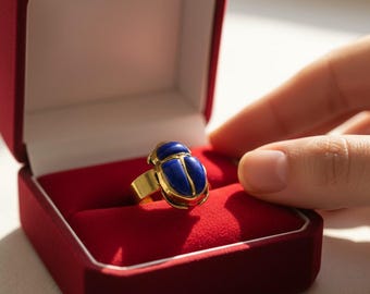 May include: A gold ring with a blue scarab beetle design, presented in a red velvet box. The ring features a detailed beetle with blue enamel and gold accents. The ring is a piece of jewelry.