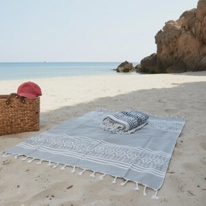 May include: A light gray beach blanket with white geometric patterns and fringe, folded on a sandy beach. A folded towel with a dark blue and white pattern rests on top. A red baseball cap and a wicker basket are nearby.