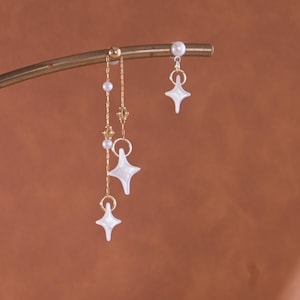 May include: Gold-toned earrings with dangling star-shaped charms and small pearl accents. The earrings are suspended from a curved bar, creating a delicate and celestial aesthetic. The star charms have a pearlescent finish.