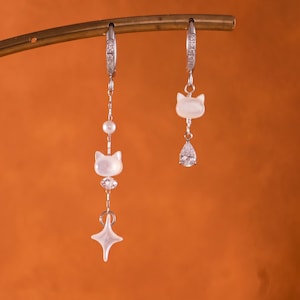 Pode incluir: Um par de brincos prateados assimétricos com pingentes em forma de gato branco. Um brinco tem uma pequena pérola, um pingente de gato e uma estrela de quatro pontas. O outro brinco apresenta um pingente de gato e um cristal transparente em forma de gota.