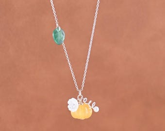 May include: A silver necklace featuring a yellow pumpkin charm with a white flower and a green leaf accent. The necklace has a delicate chain and is set against a brown background.