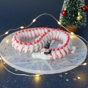 May include: A white and red dragon figurine with black horns and accents, coiled on a clear, round surface. The dragon is surrounded by fairy lights and a small Christmas tree with red and gold baubles.