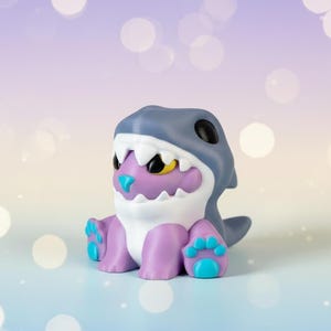 May include: A figurine of a creature wearing a shark hat. The figure is mainly purple, white, and blue, with yellow accents. The shark hat is grey, and the figure has blue paw pads.