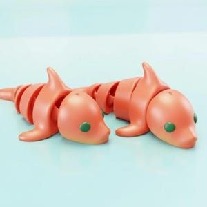 Puede incluir: Dos juguetes de delfín articulados de color naranja con ojos verdes se muestran sobre una superficie azul claro. Los delfines están conectados en forma de cadena, con cada segmento que permite el movimiento y la pose.