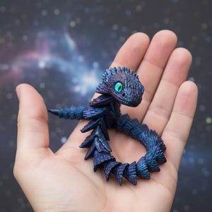 May include: A miniature, fantasy-themed sculpture of a small dragon or serpent. The creature is primarily dark blue with iridescent purple highlights, and has a detailed, textured appearance. It features a small head with a single, bright green eye.