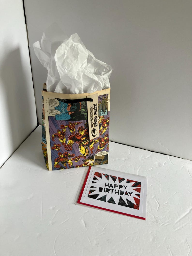 K&ouml;nnte beinhalten: Eine Geschenkverpackung mit Comic-Illustrationen und wei&szlig;em Seidenpapier. Die Tasche hat einen schwarzen Griff und den Text "Popular Designs". Eine Gru&szlig;karte mit "Happy Birthday" in einem Sternmuster ist ebenfalls vorhanden.