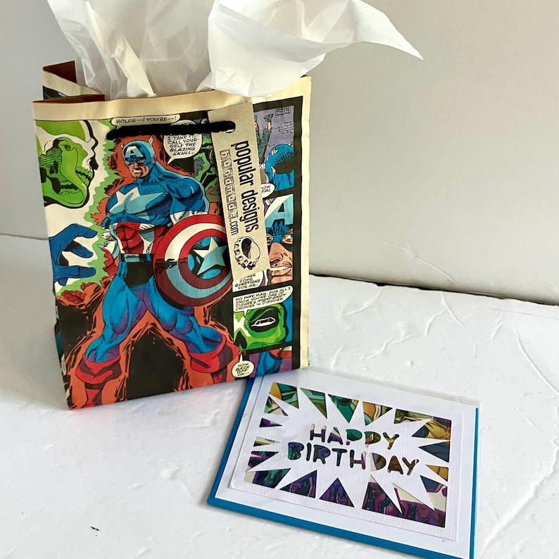 May include: A gift bag featuring a Captain America comic book print with white tissue paper inside and a tan tag labeled popular designs, accompanied by a rectangular greeting card with a white starburst cutout reading HAPPY BIRTHDAY.