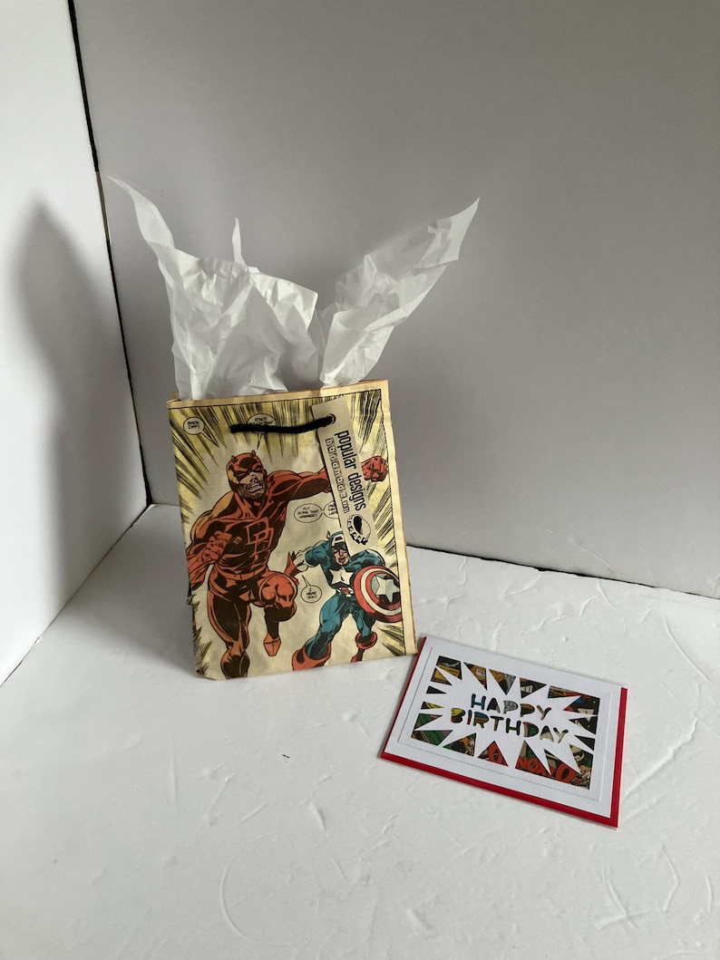 Puede incluir: Una bolsa de regalo con un dise&ntilde;o de portada de c&oacute;mic con dos superh&eacute;roes y papel de seda blanco. Una tarjeta de felicitaci&oacute;n con las palabras "Happy Birthday" est&aacute; al lado de la bolsa. La bolsa tiene las palabras "Popular Designs".