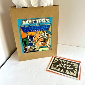 Sacola de presente e cartão comemorativo do livro Masters of the Universe