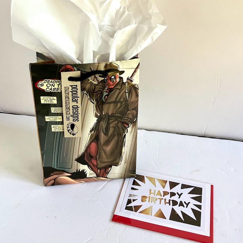 K&ouml;nnte beinhalten: Eine Geschenkt&uuml;te mit einer Comic-Illustration einer Figur im Trenchcoat und Hut mit wei&szlig;em Seidenpapier, begleitet von einer wei&szlig;en Gru&szlig;karte mit goldener Aufschrift Happy Birthday auf einer roten Kartenbasis.