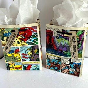 May include: Two gift bags with comic book covers featuring dinosaurs and monsters. The bags are decorated with tissue paper and tags that say "popular designs" and "handmade".