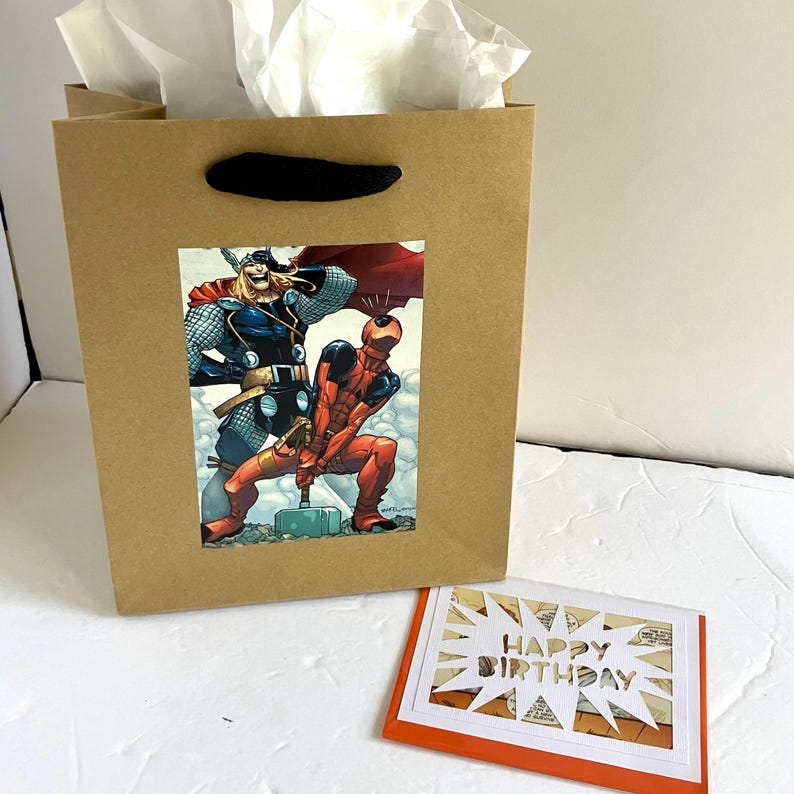 May include: A brown paper gift bag with black handles and white tissue paper, featuring a graphic of Thor and Deadpool. Beside it is a rectangular greeting card with an orange border and a white starburst cutout displaying the text HAPPY BIRTHDAY.