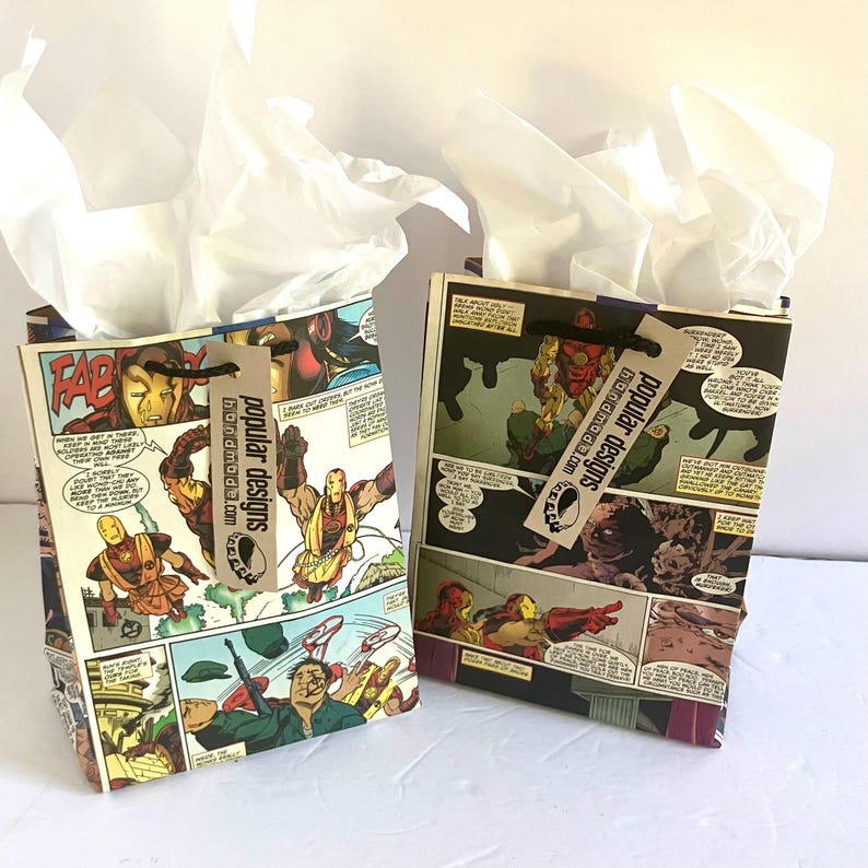 May include: Two rectangular gift bags featuring colorful comic book panels with Iron Man illustrations and white tissue paper protruding from the top. Each bag has a tan tag attached to the black handle with the text popular designs handmade.com.