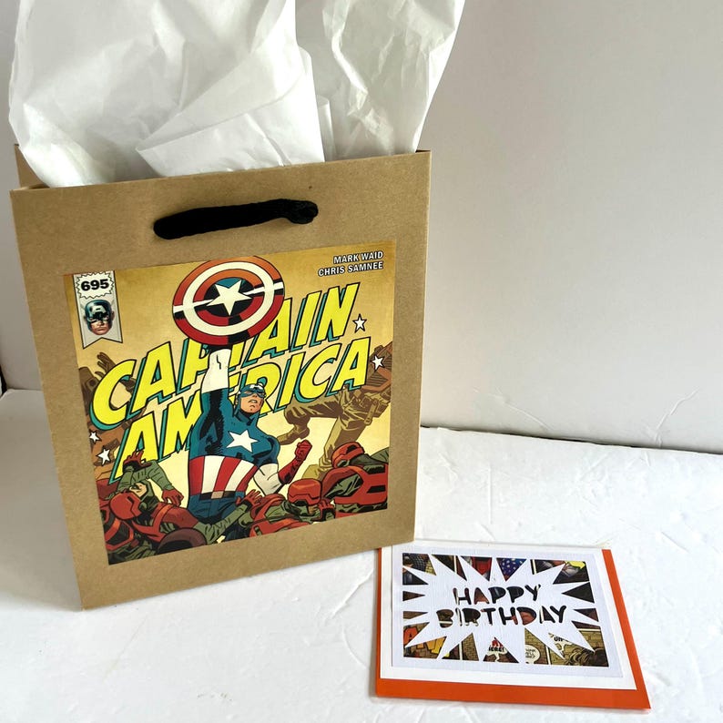 May include: A brown paper gift bag with black handles and white tissue paper, featuring a Captain America comic book cover graphic with the text Captain America, Mark Waid, and Chris Samnee. Next to it is a rectangular card with Happy Birthday text.