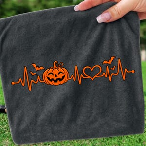 Può includere: Un capo in tessuto grigio scuro con una stampa grafica arancione brillante che raffigura una linea del battito cardiaco, una zucca di Halloween e un cuore. Piccole sagome di pipistrelli arancioni sono posizionate sopra.