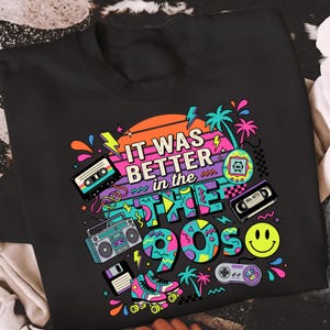 May include: A black t-shirt featuring a colorful 1990s-themed graphic print. The design includes the text "IT WAS BETTER in the 90s" surrounded by illustrations of a cassette tape, a boombox, a floppy disk, roller skates, a handheld game, a VHS tape, a smiley face, and a game controller against a sunset background.