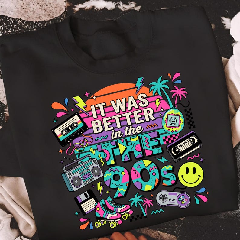 May include: A black t-shirt featuring a colorful 1990s-themed graphic print. The design includes the text "IT WAS BETTER in the 90s" surrounded by illustrations of a cassette tape, a boombox, a floppy disk, roller skates, a handheld game, a VHS tape, a smiley face, and a game controller against a sunset background.
