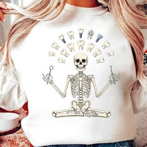 May include: White sweatshirt with a skeleton in a yoga pose, encircled by teeth and dental tools. The skeleton holds a dental mirror and a probe. The design includes various types of teeth and dental implants.