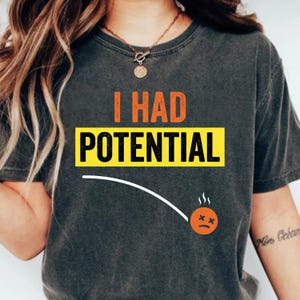 May include: Dark gray t-shirt with the text "I HAD POTENTIAL" in orange and black. The word "POTENTIAL" is in a yellow rectangle. A graphic of a sad face with steam is also on the shirt.