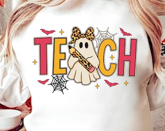 May include: White sweatshirt with a Halloween graphic. The design includes the word "TEACH" in pink and yellow block letters, a ghost holding a pencil, bats, spiderwebs, and stars. The ghost has a leopard print bow.